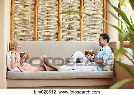 Stock Image - Couple drinking from coconuts on patio sofa. Fotosearch