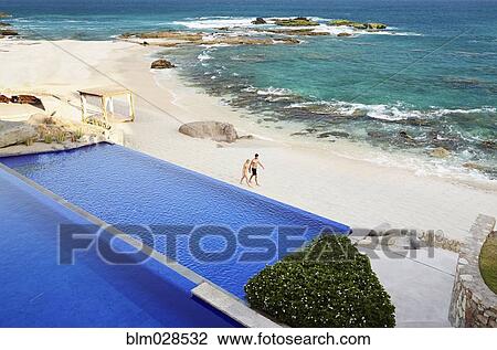 Stock Image - Couple walking on beach by infinity pool. Fotosearch