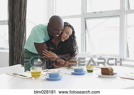 Stock Image - Couple with digital tablet hugging at breakfast table. Fotosearch