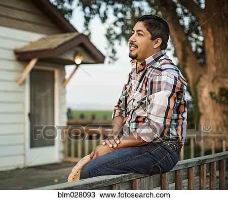 Stock Image - Hispanic man sitting on porch railing. Fotosearch