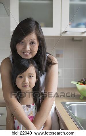 Stock Image - Asian mother hugging daughter in kitchen. Fotosearch