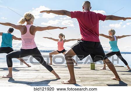 Stock Image - Caucasian people practicing yoga on wooden dock. Fotosearch