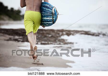 Stock Photo - Hispanic man carrying surfboard on beach. Fotosearch