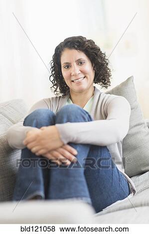 Stock Photo - Hispanic woman hugging her knees on sofa. Fotosearch