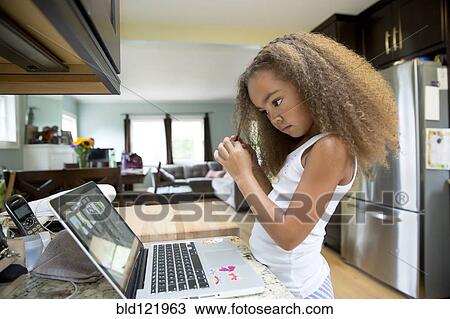 Stock Image - Mixed race girl using laptop in kitchen. Fotosearch