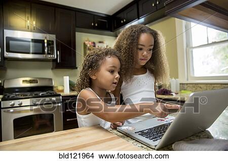 Picture - Mixed race girls using laptop in kitchen. Fotosearch