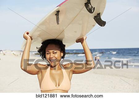 Stock Image - Mixed race woman carrying surfboard on beach. Fotosearch