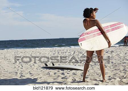 Stock Photo - Mixed race woman carrying surfboard on beach. Fotosearch