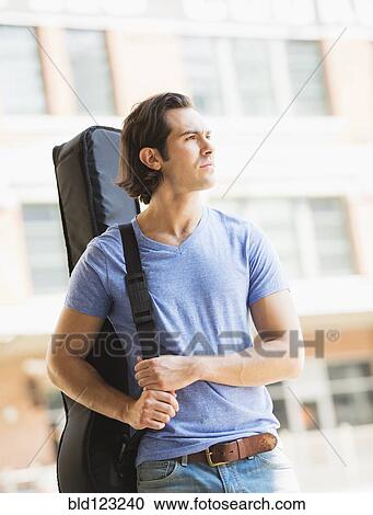 Stock Image - Caucasian man carrying guitar case on urban street. Fotosearch