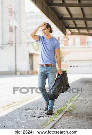 Stock Image - Caucasian man carrying guitar case on city street. Fotosearch
