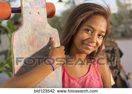 Stock Image - Mixed race teenage girl holding skateboard outdoors. Fotosearch