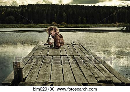Stock Image - Caucasian girl with dog on wooden deck in still lake. Fotosearch