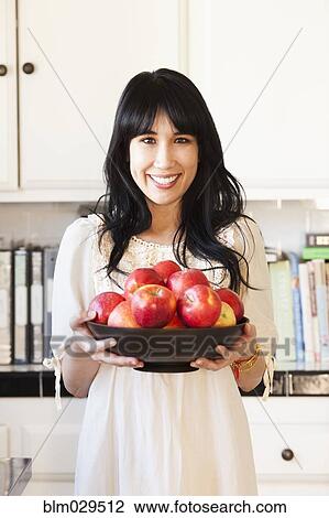 Stock Image - Woman holding bowl of fruit in kitchen. Fotosearch