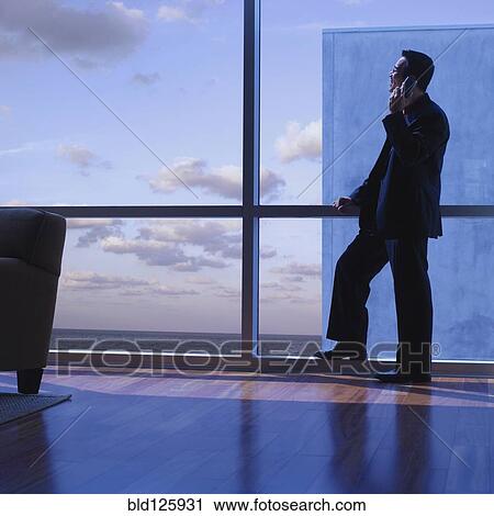 Stock Image - Asian businessman talking on cell phone at window. Fotosearch