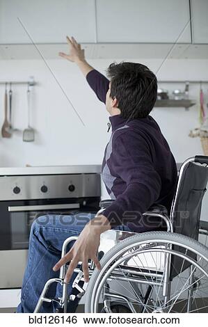 Stock Photograph - Caucasian man in wheelchair reaching for kitchen cabinet. Fotosearch