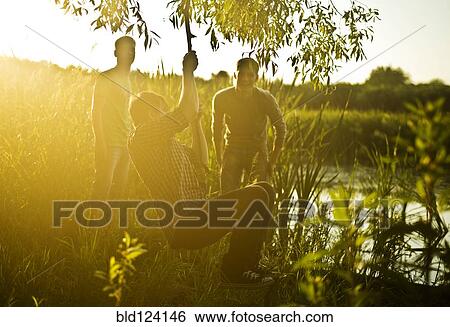 Stock Photograph - Caucasian men playing on rope swing at lake. Fotosearch