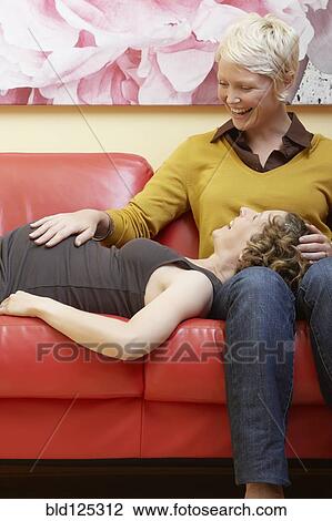 Stock Image - Caucasian pregnant lesbian couple relaxing on sofa. Fotosearch