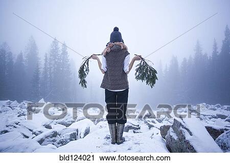 Stock Image - Caucasian woman holding tree branches on snow covered rocks. Fotosearch
