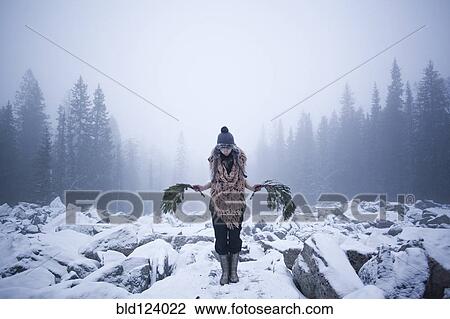 Stock Image - Caucasian woman holding tree branches on snow covered rocks. Fotosearch