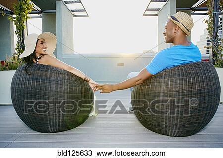 Stock Image - Couple holding hands in wicker chairs on balcony. Fotosearch