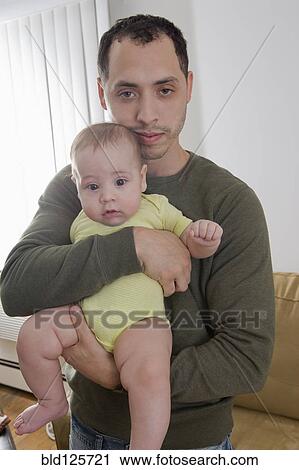 Stock Image - Father holding Hispanic baby in living room. Fotosearch