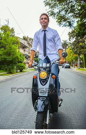 Stock Photo - Hispanic businessman standing on scooter on neighborhood road. Fotosearch