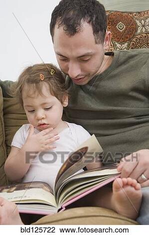 Stock Image - Hispanic father reading to daughter on sofa. Fotosearch