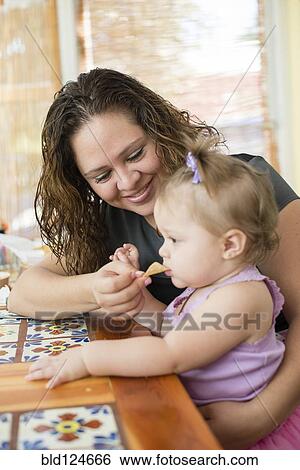 Stock Photograph - Hispanic mother feeding daughter at table. Fotosearch