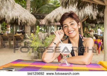 Stock Image - Hispanic woman enjoying tropical drink at resort. Fotosearch