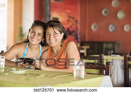 Stock Image - Hispanic women using cell phone at table. Fotosearch