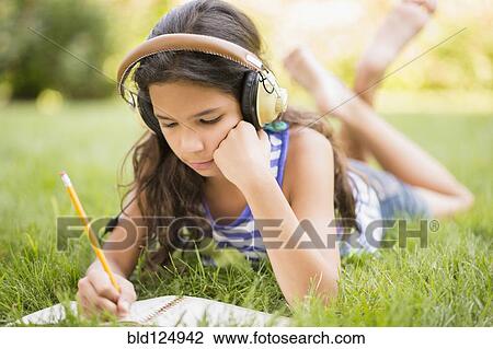 Stock Image - Mixed race girl listening to headphones and drawing in grass. Fotosearch