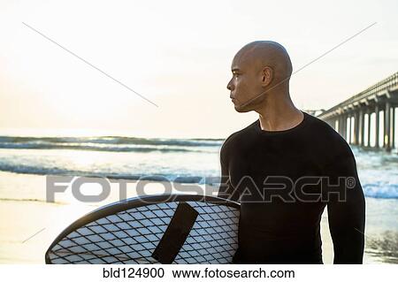 Stock Image - Mixed race man holding surfboard on beach. Fotosearch
