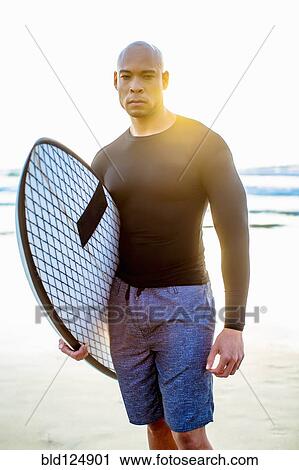 Stock Image - Mixed race man holding surfboard on beach. Fotosearch