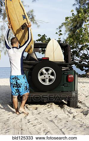 Stock Image - Mixed race man loading surfboards into 4x4 on beach. Fotosearch