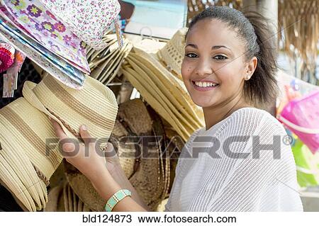 Stock Image - Mixed race woman shopping for hats at market stall. Fotosearch