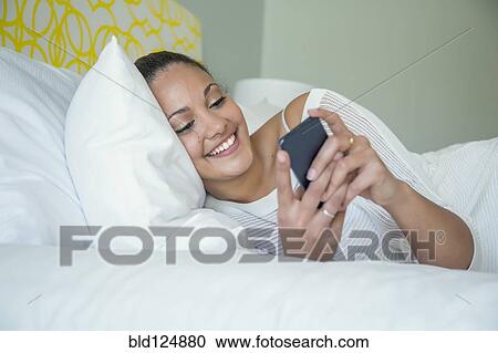 Stock Image - Mixed race woman using cell phone in bed. Fotosearch