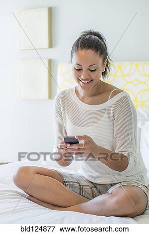 Stock Photo - Mixed race woman using cell phone on bed. Fotosearch