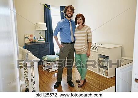 Stock Image - Pregnant Caucasian couple smiling in nursery. Fotosearch