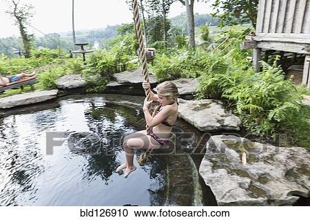 Stock Image - Caucasian girl swinging on rope above pool. Fotosearch