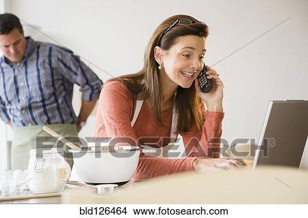 Picture - Caucasian woman baking, using laptop and talking on telephone. Fotosearch