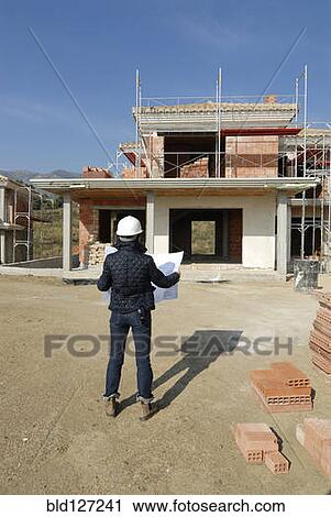 Stock Image - Female architect on construction site looking at plan rear view. Fotosearch