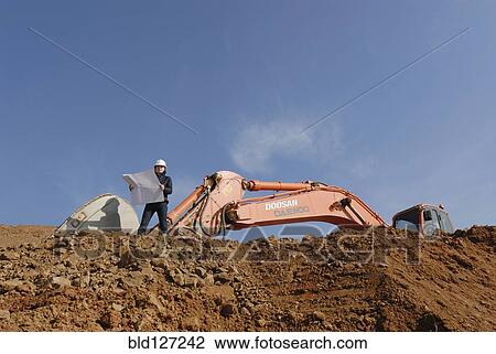 Female engineer holding blueprint standing in front of excavator low angle view View Large Photo Image Stock Image - Female engineer holding blueprint standing in front of excavator low angle view. Fotosearch