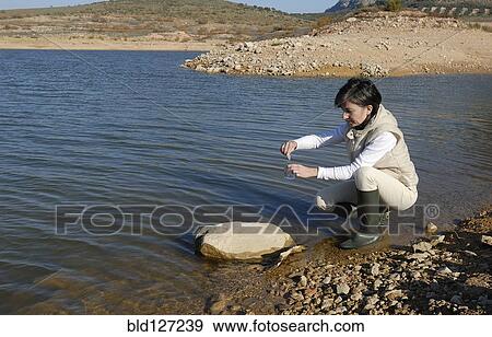 Stock Photo - Female scientist taking water sample from lake side view. Fotosearch