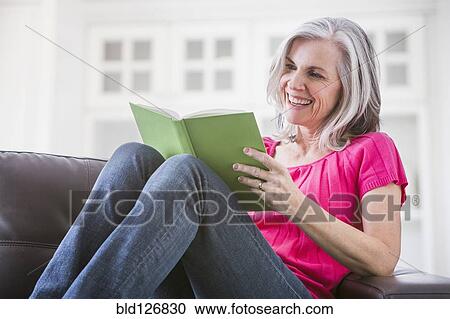 Stock Image - Happy Caucasian woman reading book on sofa. Fotosearch