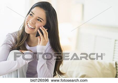 Stock Photograph - Happy Hispanic woman talking on cell phone in bed. Fotosearch
