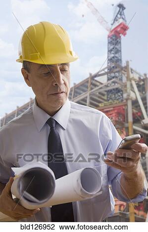 Stock Image - Hispanic architect using cell phone at construction site. Fotosearch