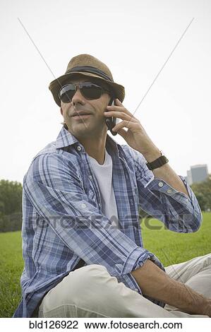 Stock Image - Hispanic man talking on cell phone in grass at park. Fotosearch