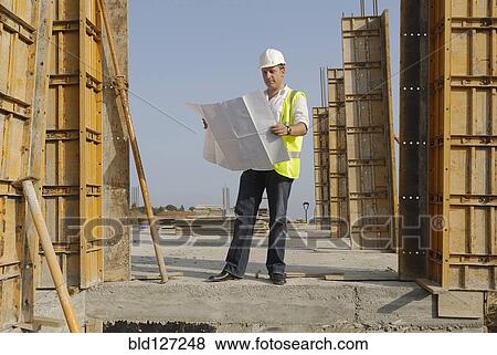 Male engineer wearing hard hat looking at plans on building site View Large Photo Image Stock Photo - Male engineer wearing hard hat looking at plans on building site. Fotosearch