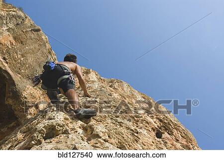 Stock Image - Male free climber scaling rock face rear view upward view. Fotosearch