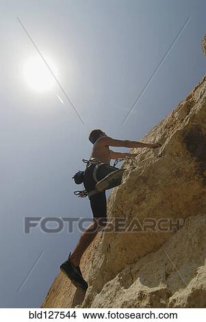 Picture - Male rock climber scaling rock face secured by ropes low angle view. Fotosearch